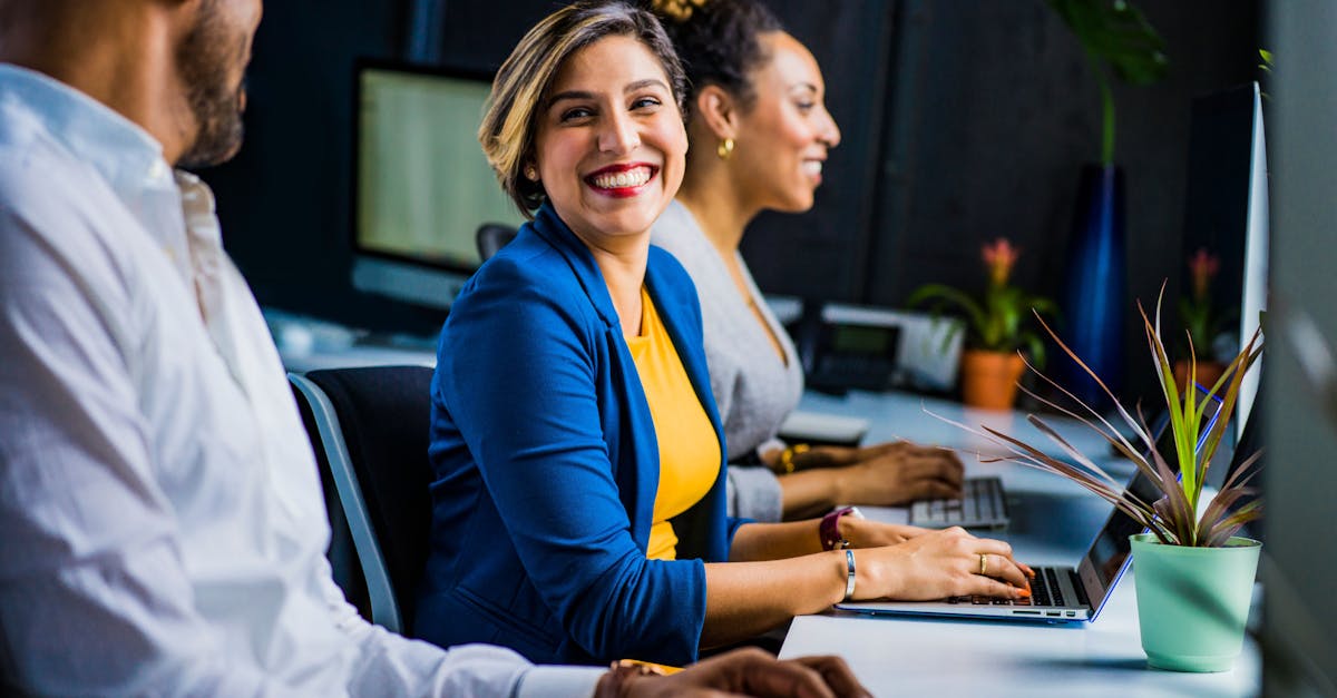 Three diverse professionals working and smiling at office desks, fostering teamwork and collaboration.
