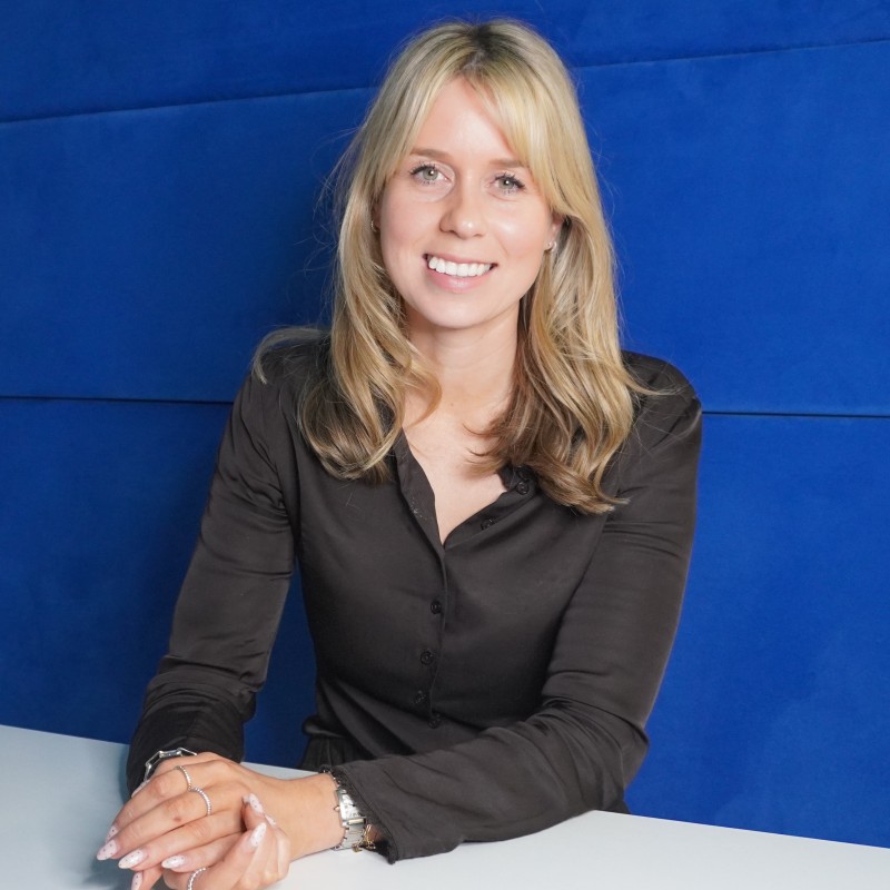 Aimee Doole - Industry 5.0 - Industry 5.0 Smiling blonde woman in a dark blouse sits at a white table against a blue background, facing the camera.