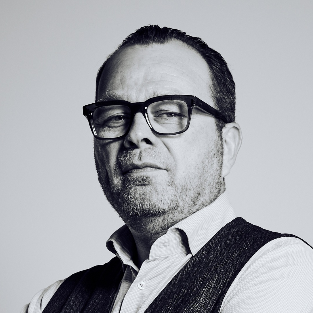 Portrait of a middle-aged man with dark hair and glasses, wearing a light shirt and dark vest.