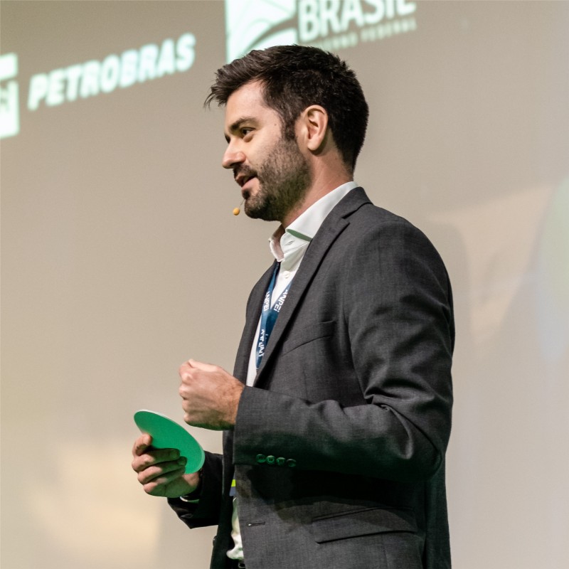 Man in a dark suit giving a talk on stage, holding a green circular card with sponsor logos visible in the background.