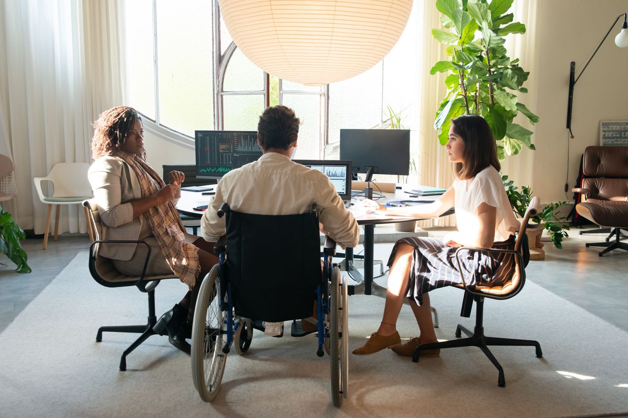 Team meeting in bright office with three people around a desk; one person in a wheelchair discusses with two colleagues, monitors showing charts.