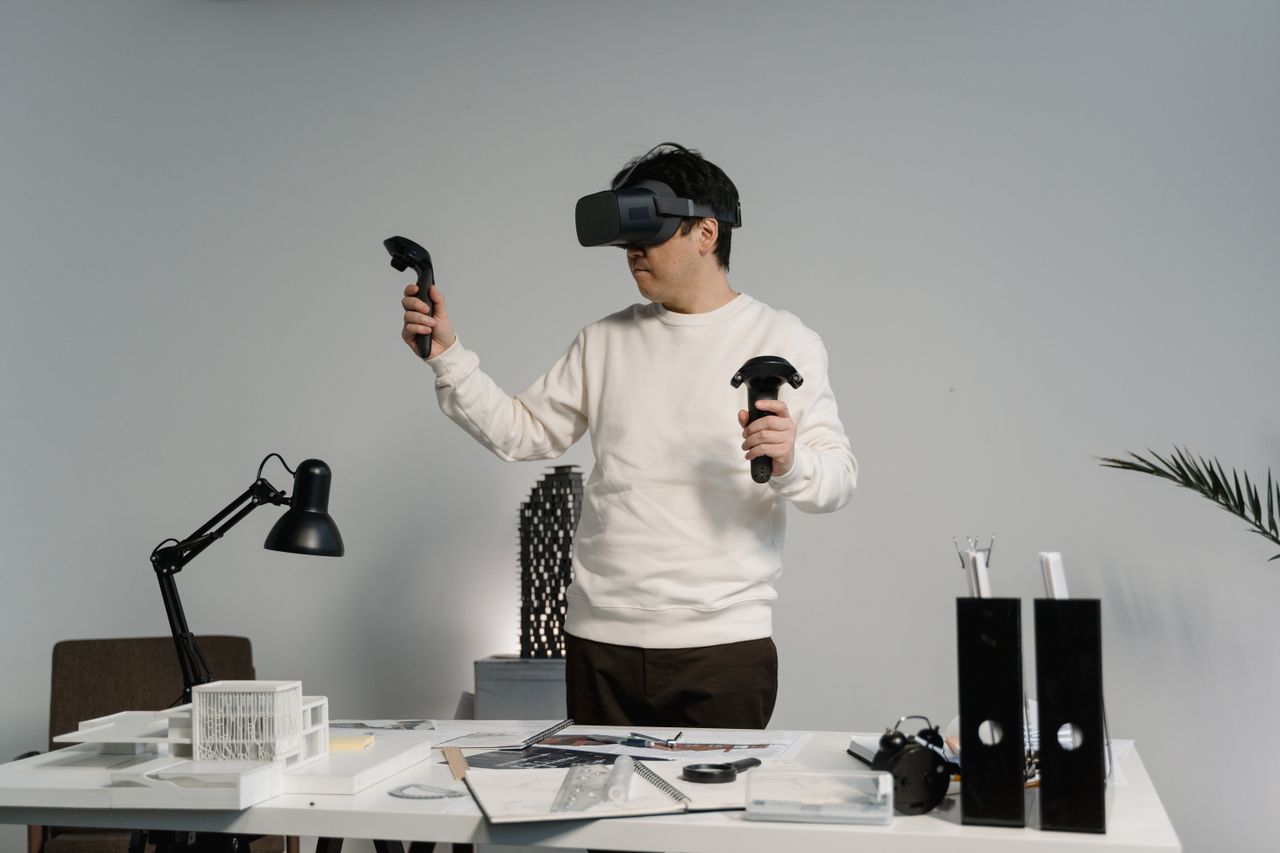 Man wearing a VR headset stands at a desk, holding two motion controllers, immersed in virtual reality setup.